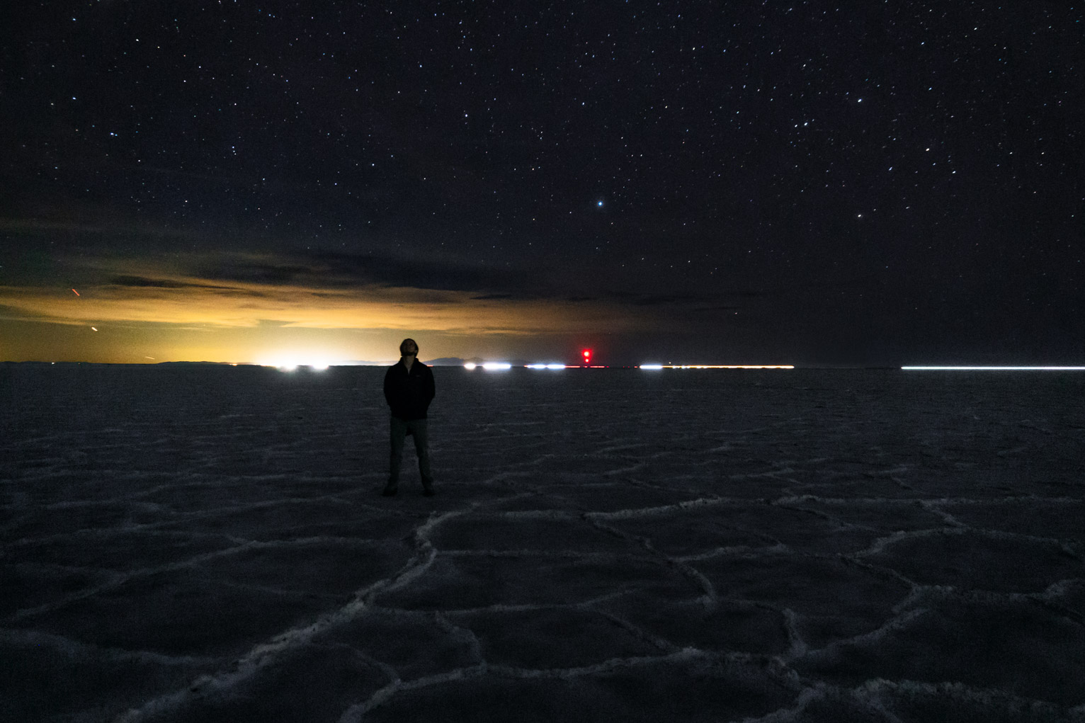 on the horizon a group of clouds is lit bright yellow by, I believe, a military base, above the starry dark blue sky, and below the textured salt flats